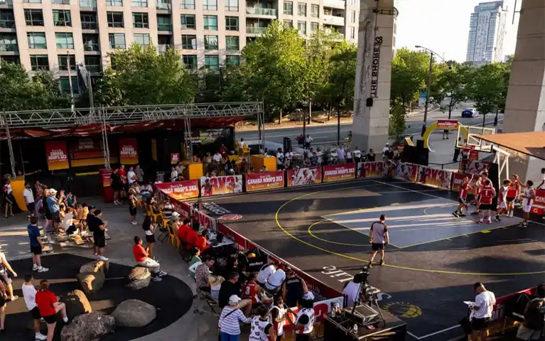 Wide angle view of outdoor basketball court at The Bentway showing spectators watching Sun Life Canada Hoops 3x3 tournament in downtown Toronto urban setting with championship court branding