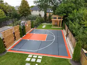 An expansive 30x40 backyard basketball court featuring premium interlocking tiles in slate and orange. This Mississauga installation includes a professional hoop, custom fencing, and integrated landscaping, creating a high-end athletic space for the whole family.
