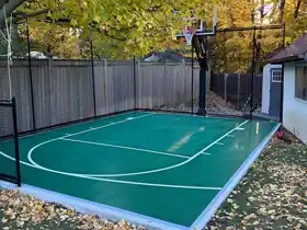 A space-efficient 18x25 basketball court in Scarborough featuring a vibrant green surface and perimeter netting. This professional installation maximizes a smaller backyard footprint, providing a dedicated high-performance practice area framed by beautiful natural landscaping.