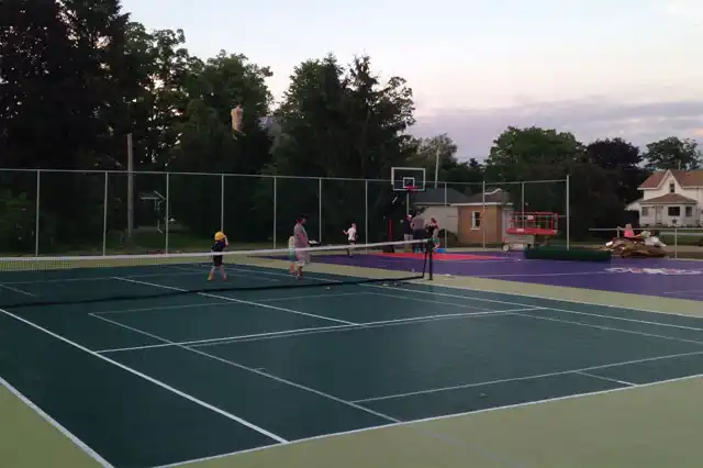 Public outdoor multi-sport court with a green tennis court in use and a purple basketball half-court in the background, surrounded by fencing and residential homes.